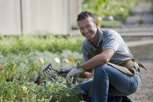 A man with short dark hair and a friendly expression is kneeling in a garden, holding a small garden rake in his right hand while tending to a variety of green leafy plants and flowers. The garden features a well-maintained lawn with dense, lush grass and neatly edged flower beds filled with small plants and blooming flowers, possibly pansies or similar. In the background, there is a wooden fence, and the garden appears to be in a residential outdoor space, with natural daylight illuminating the scene on a clear day. The man is dressed in a light blue shirt, jeans, and a gardening apron, suggesting he is engaged in lawn or garden maintenance services. This setting likely represents a typical front or backyard in the Plaistow area, where professional gardening services such as those offered by Lawn Mowing Plaistow could be applicable.